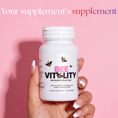 A hand with white nail polish holds a bottle labeled Bee Vitality 🐝 against a pink background. Text above reads, Your supplement’s supplement, showcasing the power of royal jelly and fermented bee bread.