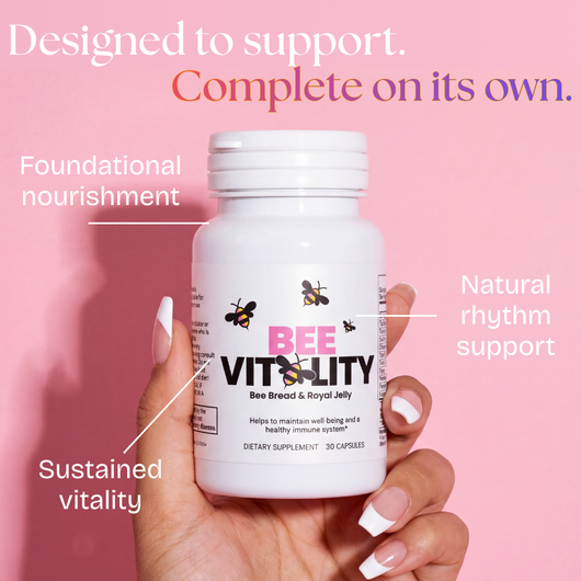 A hand with white-painted nails holds a bottle of Bee Vitality 🐝 capsules against a pink background, highlighting benefits like foundational nourishment, sustained vitality, and support for your natural rhythm.